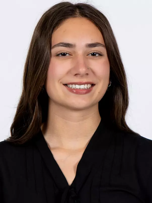 Headshot of Kailyn Tibolla smiling, wearing a black v-neck blouse with small hoop earrings and long dark brown hair.