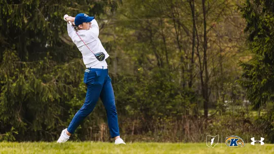 A Kent State women's golfer completes a full follow-through on a driver swing, wearing a white long-sleeve top, blue pants, white shoes, and a blue Kent State cap, with a wooded course backdrop and the Kent State lightning bolt, Golden Flashes, and Under Armour logos in the lower right corner.