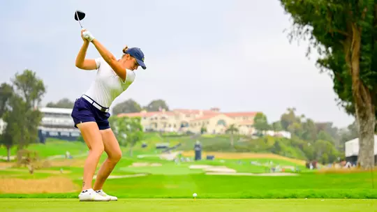 veronika kedVeronika Kedronova winds up at the top of her driver swing on the tee, wearing a white short-sleeve top, navy shorts, white Fila shoes, and a navy Kent State cap, with the ball on the tee in front of her, a lush tournament course, and a Spanish-style clubhouse visible in the background.onova at US Open