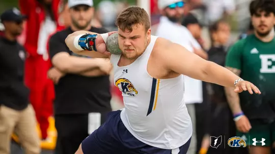A Kent State track and field athlete competing in the shot put, wearing a white Under Armour singlet with the Kent State Golden Flashes logo and navy shorts. He grips the shot put against his neck with a taped right hand and braced wrist, his left arm extended back for balance during the throwing motion, with an intense, focused expression. Coaches, athletes, and spectators are visible blurred in the background, including an Eastern Michigan athlete in a green jersey. The Ohio state outline, Kent State Golden Flashes logo, and Under Armour logo appear in the bottom right corner.