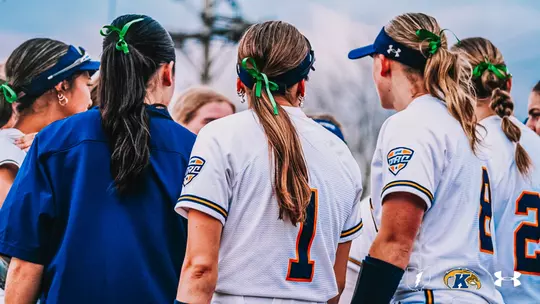 A close-up photo taken from behind of several Kent State softball players gathered in a huddle on the field during a game. The players are wearing white Kent State home uniforms with navy and gold trim on the sleeves and MAC Conference shield patches on the right shoulder. Jersey number 1 is clearly visible in navy with an orange outline on the center player, and a partial jersey number is visible on the right. All players visible are wearing green ribbon hair ties or accessories, with their hair pulled back in ponytails or braids. A coach or staff member in a navy blue jacket is partially visible on the left side of the frame. One player on the right is wearing a black arm sleeve. The background shows a softly blurred outdoor softball field and overcast sky. The Ohio state outline with lightning bolt, Kent State Golden Flashes logo, and Under Armour logo are displayed in the bottom right corner of the image.