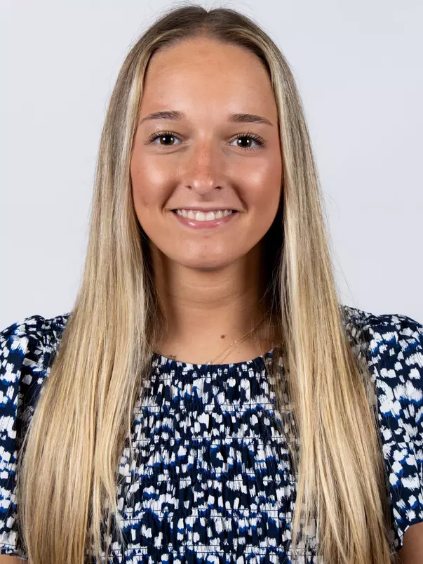 Headshot of Emma Masse smiling, wearing a navy and white patterned short-sleeve top with a delicate gold necklace, and long straight blonde hair.