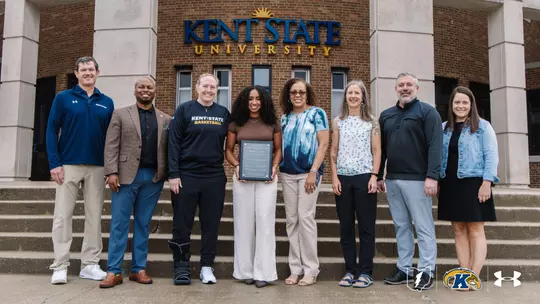 Dionna Gray holds an award plaque while standing with seven Kent State University administrators, coaches, and staff members on the steps in front of Kent State University's building entrance. A Kent State women's basketball coach stands to her left wearing a walking boot.