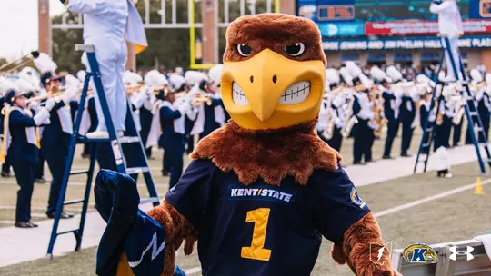Flash, the Kent State Golden Flashes mascot, stands on the sideline in a navy 'Kent State' No. 1 football jersey, with the Kent State marching band performing in white and navy uniforms in the background and the Kent State lightning bolt, Golden Flashes, and Under Armour logos in the lower right corner.