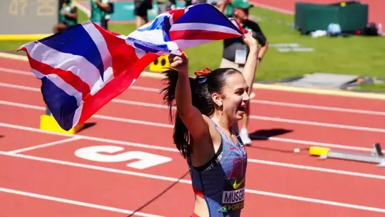 A track and field athlete wearing a Great Britain uniform celebrates while running with arms raised, waving a large Union Jack flag overhead. She has a dark ponytail with a red ribbon and is smiling with her mouth open in celebration. The scene is set on a red track with white lane markings, with a scoreboard and stadium officials visible in the blurred background.