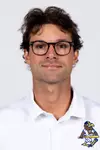 Studio headshot of a young man with dark wavy hair and dark-rimmed glasses, wearing a white Kent State baseball polo shirt against a plain light gray backdrop.