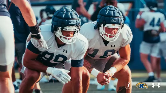 Two Kent State football players crouch side-by-side in a low stance during practice, wearing white practice jerseys and padded Guardian caps on their helmets.