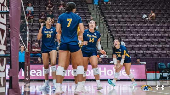 Four Kent State volleyball players in navy blue jerseys celebrate on the court together during a match, with maroon arena seating and a "Health Care for Hokies" sponsor board visible in the background.