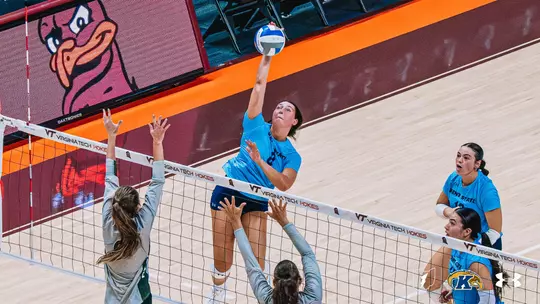 A Kent State volleyball player in a light blue jersey jumps to spike the ball over the net during a match against William & Mary. Two William & Mary blockers in gray jerseys reach up to defend at the net, while two Kent State teammates in blue jerseys look on from the right side of the court. The Virginia Tech Hokies mascot is displayed on a video board in the background.