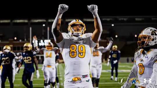 Kent State Golden Flashes player wearing No. 89 raises both fists in celebration on the field during a night road game, in a white road uniform with gold accents and a white helmet with gold facemask, with teammates and opposing players visible in the background and the Kent State lightning bolt, Golden Flashes, and Under Armour logos in the lower right corner.