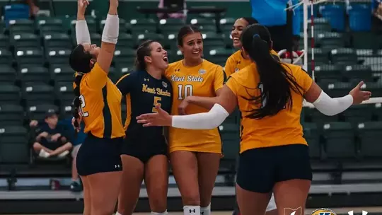 Five Kent State volleyball players celebrate on the court, wearing a mix of navy and gold jerseys, with arms raised and smiling. A referee in a blue shirt stands near the net in the background, with empty green stadium seating behind them.