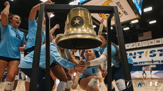 Kent State volleyball players in light blue jerseys celebrate around the "Starner Victory Bell" after a win, with two players ringing the bell by its rope inside M.A.C. Center, Kent State banners and an American flag visible in the background.