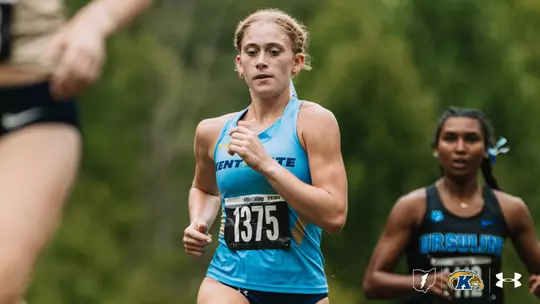 A Kent State cross country runner wearing bib No. 1375 competes in a race, running past a blurred competitor in the foreground. An Ursuline runner is visible behind her on the course.
