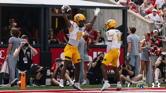 A Kent State football player wearing jersey number 18 leaps into the air with both arms raised holding a football, celebrating an interception in the end zone alongside a teammate wearing number 14.