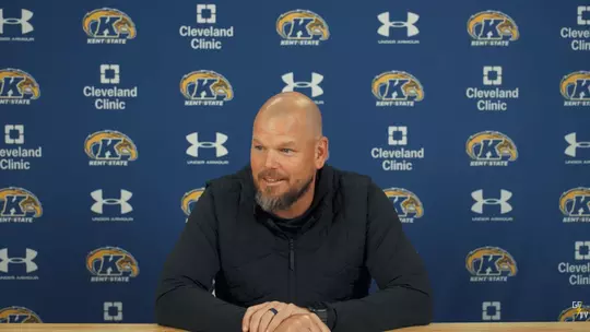 A smiling man with a beard sits at a wooden table during a press conference in front of a blue Kent State backdrop.