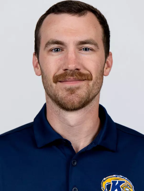 Headshot of a smiling man with short dark hair, a mustache, and facial stubble wearing a navy blue Kent State polo shirt against a plain background.