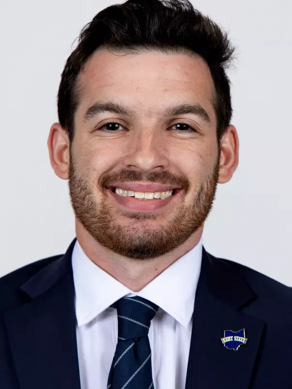 Headshot of a smiling man with short dark hair and a beard wearing a dark suit, tie, and a Kent State lapel pin against a plain background.