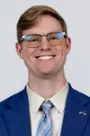 Headshot of a smiling man with short light brown hair and glasses wearing a blue suit jacket, striped tie, and a Kent State lapel pin against a plain background.