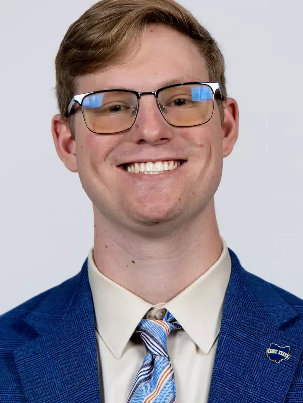 Headshot of a smiling man with short light brown hair and glasses wearing a blue suit jacket, striped tie, and a Kent State lapel pin against a plain background.