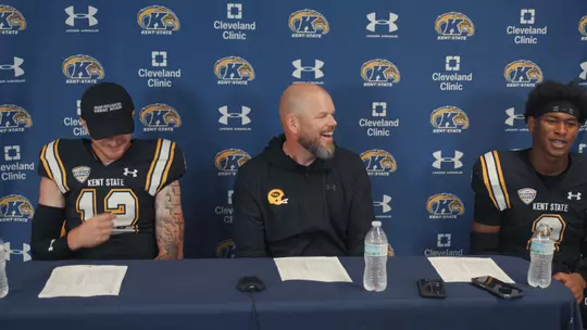Press conference scene featuring two Kent State football players and a coach seated at a table in front of a branded media backdrop, smiling and talking