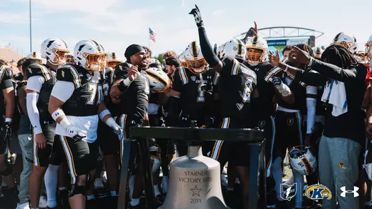 Kent State football players gathering in celebration around the Starner Victory Bell on the field.