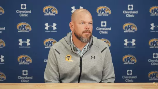 A bald coach with a beard wearing a gray Kent State hooded jacket seated in front of a branded media backdrop.