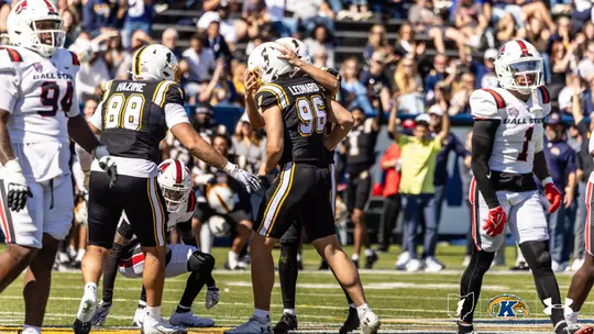 Action photo of Kent State football players embracing in celebration while opposing Ball State players stand nearby on the field.