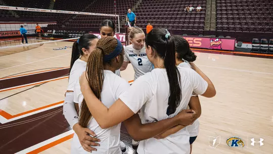 Kent State volleyball players huddle together with arms around each other's shoulders before a match at Virginia Tech's arena, with the Hokies' logo and empty maroon seating visible in the background. Player No. 2 stands in the center of the huddle wearing a white Kent State jersey.