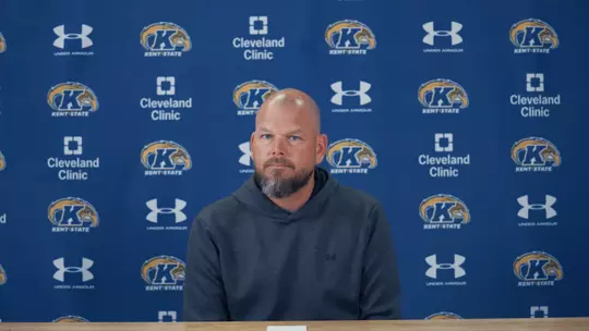 A coach sitting at a press conference table in front of a blue Kent State step-and-repeat backdrop wearing a dark gray hoodie.