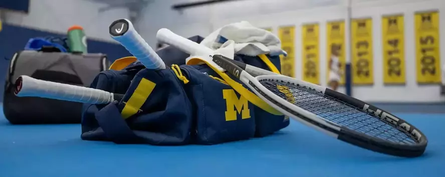 Tennis bag on the court with raquet on top of the bag and Michigan banners in the background