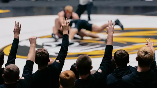 A group of teammates in black warmups stand at the edge of a wrestling mat with their backs to the camera, raising three fingers in the air as they react to the action. In the blurred background, two wrestlers are on the mat during a match while a referee kneels nearby, with a crowd of spectators seated behind them in the arena.