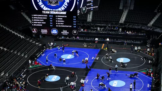 Wide overhead view of a large indoor arena set up for the NCAA wrestling championships, with four wrestling mats on the floor where athletes and coaches are warming up and drilling. A massive video board above the mats displays “NCAA Championships” and team standings, while mostly empty arena seats surround the competition area and event staff sit at tables along the edge of the mats.
