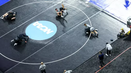 Overhead view of multiple wrestlers warming up on a large black mat at an NCAA venue. Several pairs grapple in different areas of the circle, while coaches and staff stand along the edge observing. Gear and equipment sit near the boundary, and a scoreboard is visible off to the side.