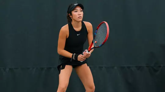 A focused tennis player in a black athletic outfit and visor stands in a ready position on the court, gripping a red and black racket with both hands. She faces forward with an alert expression, knees slightly bent, prepared to receive a shot. The background is a dark, plain court barrier, emphasizing her stance and concentration.