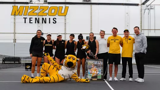 A group of University of Missouri women's tennis players and coaches stand together on an indoor court beneath a large “MIZZOU TENNIS” sign. The team, dressed mostly in black and gold, surrounds a senior player holding flowers, while a framed photo rests on the ground in front. The school's tiger mascot lounges playfully at the front of the group.