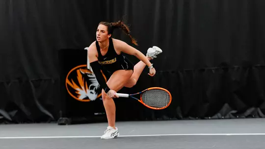 A female tennis player in a black Mizzou uniform lunges forward mid-swing on an indoor court, her body low and balanced as she tracks the ball. She grips a bright orange racket with both hands, her hair pulled back in a ponytail, and wears white tennis shoes. A large tiger logo and dark curtain backdrop fill the background, emphasizing the intensity and focus of the moment.
