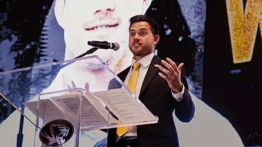 Robin Goodman in a black suit and gold tie speaks at a clear podium with a Mizzou Tigers logo, gesturing with one hand while standing in front of a large screen displaying a close-up portrait behind him. A microphone is positioned at the podium during the indoor event.