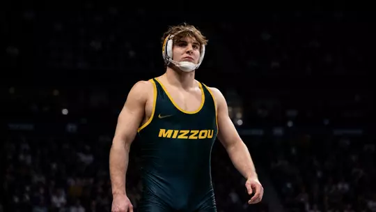 Luke Geog wearing a black and gold “MIZZOU” singlet and white headgear stands under arena lighting, looking focused and determined. He is centered in the frame with a strong, upright posture, while a dark, blurred crowd fills the background, creating a dramatic, spotlight effect around him.