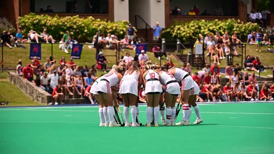 Spider field hockey in their all-white uniforms with red numbers huddles prior to the start of the game.
