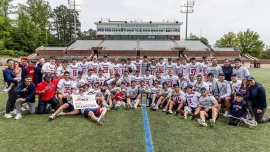 Richmond men's lacrosse team huddles around the 2026 Atlantic 10 Men's Lacrosse Championship trphiy