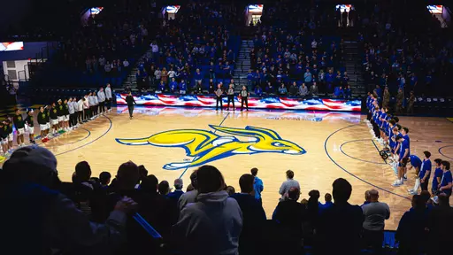A basketball arena filled with spectators stands for a pregame ceremony. Two teams line up on opposite sidelines of the court, which displays a large yellow-and-blue jackrabbit logo at center. A long digital board along the scorer's table shows an American flag graphic.