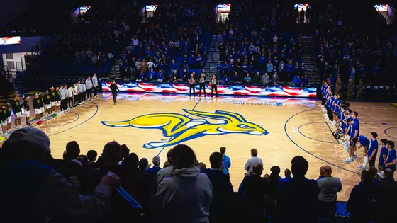 A basketball arena filled with spectators stands for a pregame ceremony. Two teams line up on opposite sidelines of the court, which displays a large yellow-and-blue jackrabbit logo at center. A long digital board along the scorer's table shows an American flag graphic.