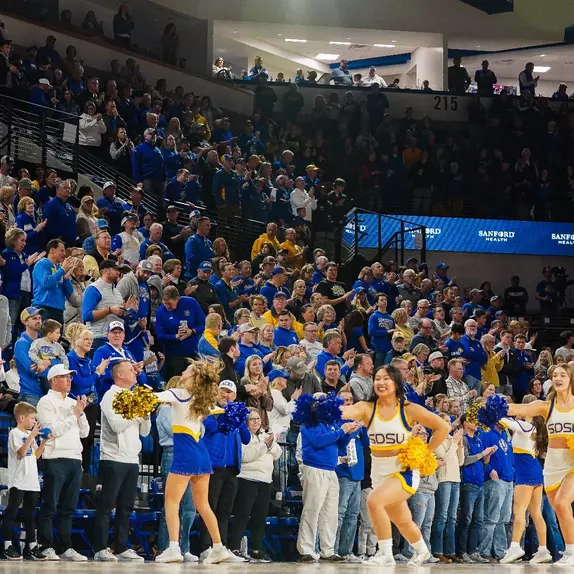 Cheerleaders in white uniforms on the court while fans in the stands in yellow and blue attire applaud