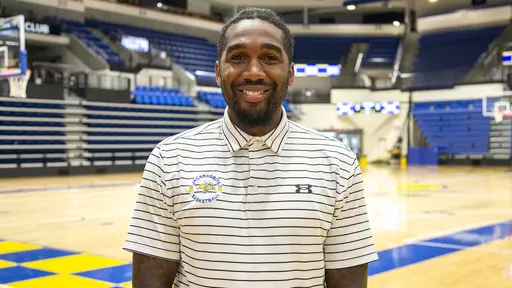 A picture of a man in a white polo with black stripes smiling posed inside a basketball arena