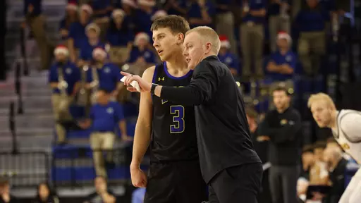 A coach in a black long sleeve jacket instructs a player beside him while the basketball player in a black uniform listens