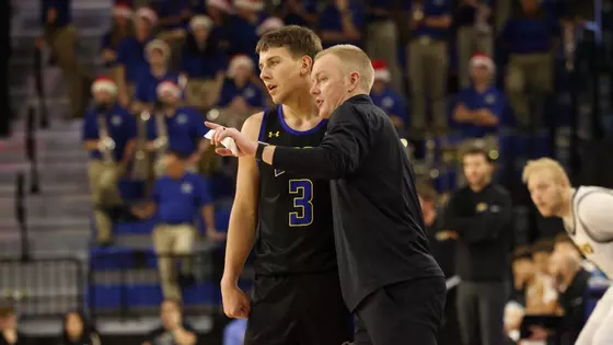 A coach in a black long sleeve jacket instructs a player beside him while the basketball player in a black uniform listens