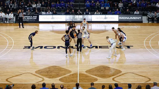 Ten men's basketball players, with one team in dark blue and another in white uniforms, stand ready on a court prior to tipoff of a game
