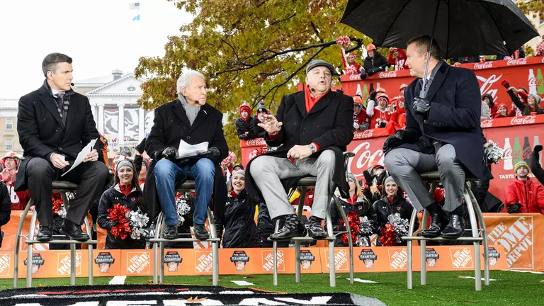 As cold rain falls and Wisconsin Badger and Michigan Wolverine fans wave signs and cheer in the background, Wisconsin Athletic Director Barry Alvarez, second from left, joins ESPN College GameDay commentators on stage during a live broadcast of the show on Bascom Hill at the University of Wisconsin-Madison on Nov. 18, 2017. Immediately after the show, the Badgers play Michigan in a football game at Camp Randall Stadium. (Photo by Jeff Miller / UW-Madison)