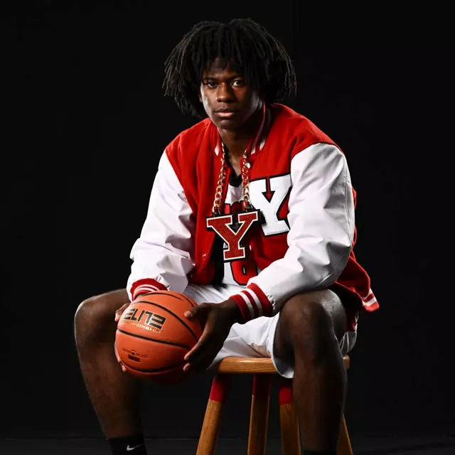Men's basketball student-athlete Jaiden Haynes sits on a stool holding a basketball and wearing his uniform, a YSU letterman's jacket and a Y chain in the digital media studio.