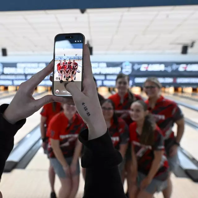 A cell phone is in focus as assistant bowling coach Madyson Marx uses it to take a photo of the 2026 YSU bowling team with lanes in the background.